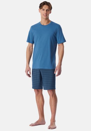 Ung mand, der står barfodet, iført en enkel blå kortærmet skjorte og blå mønstrede shorts mod en hvid baggrund.