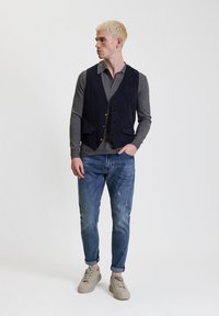 Marineblauer Weste über einem grauen Langarmshirt, kombiniert mit verwaschenen blauen Jeans und beigen Sneakers. Glatter Stoff, vier Taschen, Knopfdetails.