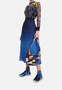 Osoba ubrana w kolorową bluzkę z długim rękawem w kwiaty, niebieską plisowaną spódnicę, niebieskie skarpetki oraz błyszczące trampki z Myszką Miki, trzymająca torebkę w kwiatowy wzór.