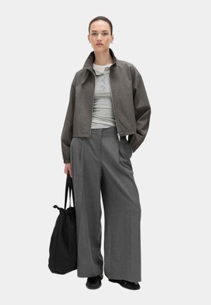 Kvinde stående iført grå kort jakke, lysegrå henley-trøje, grå bukser med brede ben, sorte sko og holder en stor sort tote taske.