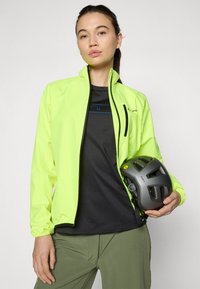 Neón žltá ľahká bunda s zipsovými vreckami, spárovaná s čiernym tričkom a olivovozelenými nohavicami. Model drží šedú cyklistickú prilbu.