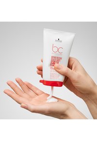 Crème Bonacure Repair Rescue dans un tube blanc avec des accents rouges. Produit blanc crémeux est distribué sur une main. 100 ml.
