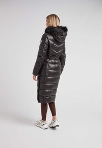 Abrigo de plumas negro con capucha y ribete de pelo sintético, que presenta una cintura ceñida con cinturón y un diseño panelado. Llevado sobre leggings marrones y zapatillas.