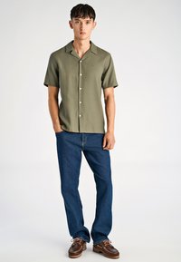 Młody mężczyzna stoi z rękami w kieszeniach, ubrany w oliwkową koszulę na krótki rękaw zapinaną na guziki, niebieskie dżinsy i brązowe skórzane buty.