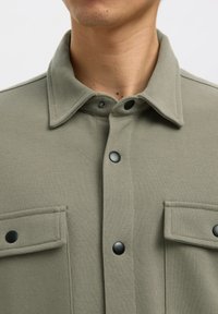 Hombre con una chaqueta camisa verde caqui abotonada con dos bolsillos delanteros con solapa y cierres de presión negros.