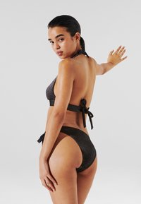 Svart, skimrende bikini med halter-top og knyttet bak. Bunndelen har et høyt snitt og minimal dekning, som fremhever strukturert stoff.