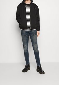 Svart bomberjacka med dragkedja framtill, randig T-shirt, slitna blå jeans och svarta ankelstövlar med gula sömmar.