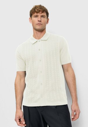 Mann mit lockigem Haar, der ein kurzärmeliges, weißes Strickhemd mit Knöpfen und schwarze Hose trägt und vor einem einfachen hellen Hintergrund steht.