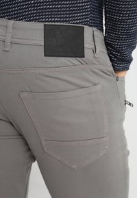 Graue Baumwollhose mit glatter Textur, ausgestattet mit einem schwarzen Lederschild und einer seitlichen Reißverschlusstasche. Klassisches Fünf-Taschen-Design.