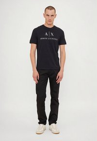 Must lühikeste varrukatega T-särk, millel on valge "A|X" logo ja "ARMANI EXCHANGE" tekst, kombineeritud mustade pükste ja valgete tossudega. Lihtne ja vormis lõige.