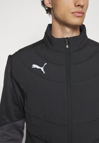 Schwarze Jacke mit Reißverschluss, hohem Kragen und strukturierten Einsätzen. Weißes Logo auf der Brust. Mit grauen Ärmeln und leichtem Material.