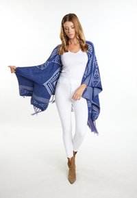 Poncho albastru cu un model geometric, cu franjuri, țesut dintr-un material moale. Are un design geometric și un decolteu deschis, purtat peste un top alb.