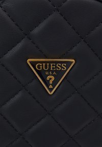 Sac à main en cuir matelassé noir avec un logo triangulaire doré orné d'accents noirs, affichant "GUESS U.S.A." et un emblème de point d'interrogation.