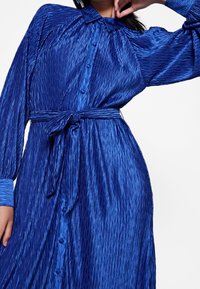 Robe plissée bleue en tissu brillant. Présente un devant à boutons, des manches longues et une ceinture nouée à la taille.