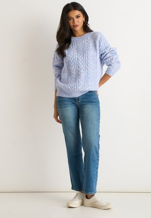 Woman standing with hand on hip wearing a light blue cable knit sweater, straight-leg blue jeans, and white sneakers against a plain background.