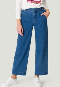 Jean denim bleu à jambes larges avec une taille haute, doté de poches latérales et d'une finition nette. Associé à des baskets blanches pour un look décontracté.