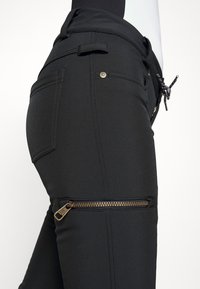 Svarta stretchbyxor med en slät textur, med en siddragkedja och guldfärgade detaljer. Ytterligare fickor och justerbar midjedetalj.