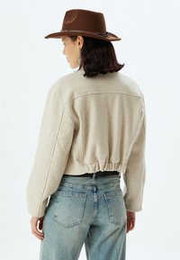 Maglione beige corto con orlo elasticizzato, maniche lunghe e vestibilità rilassata, abbinato a jeans azzurri a vita alta e un cappello marrone.