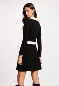 Vestido negro con mangas largas, cuello blanco y bodice ajustado que se amplía en una falda. Incluye una pulsera de puño dorada. Textura suave.