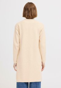 Manteau long beige à coupe décontractée, fabriqué en tissu texturé. Présente des manches longues et un ourlet droit, associé à un jean large bleu.