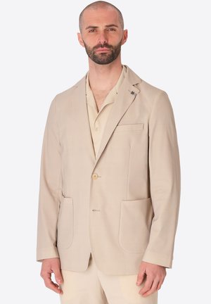 Uomo con barba che indossa una giacca beige con grandi tasche anteriori e una camicia coordinata con colletto, in piedi di fronte.