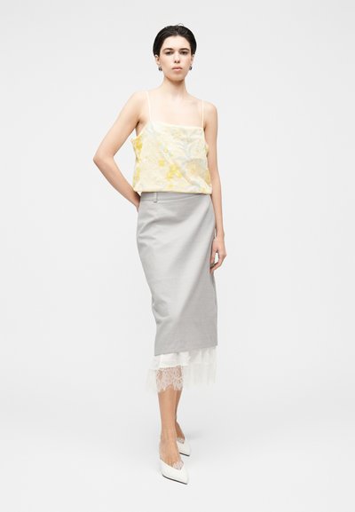 Κίτρινο κεντητό crop top με λεπτές τιράντες, σε συνδυασμό με φούστα pencil σε ανοιχτό γκρι χρώμα και δαντέλα στο τελείωμα. Το μοντέλο φοράει λευκά μυτερά παπούτσια.