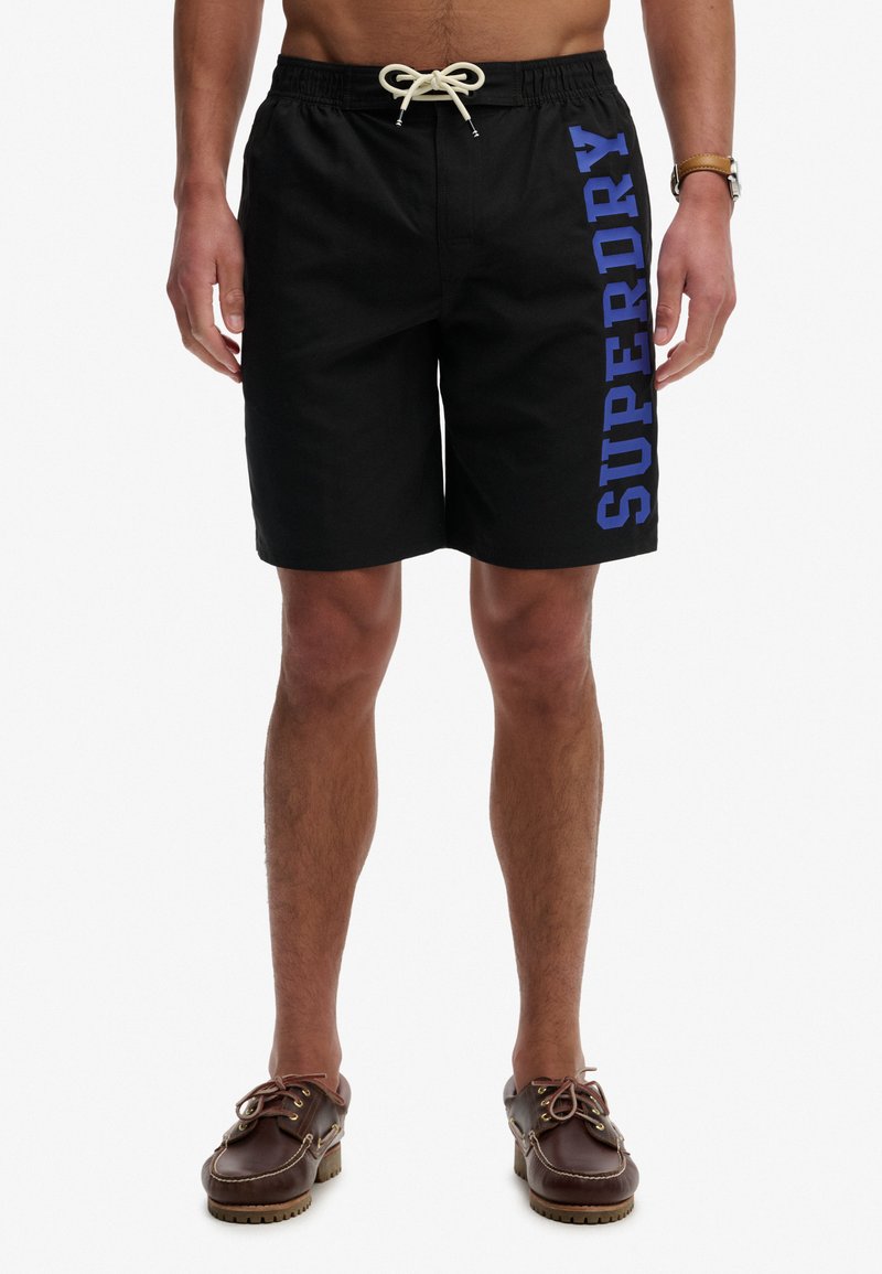 Homme portant un short de bain noir Superdry avec un cordon blanc et des chaussures bateau en cuir marron, se tenant devant un fond blanc.