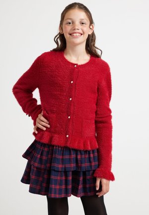 Rode harige cardigan met een ronde halslijn, knopen aan de voorkant en een gerimpelde onderrand. Gecombineerd met een marineblauw geruit gelaagdrok en zwarte panty’s.