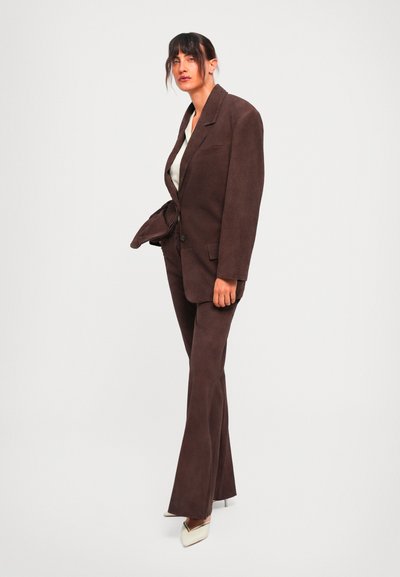 Žena u tamnosmeđem oversized blejzeru i odgovarajućim širokim pantalonama, sa belim cipelama na štiklu, stoji naspram jednostavne svetle pozadine.