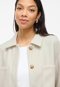 Ανοιχτό μπεζ μπουφάν από μείγμα μαλλιού με κολάρο, δύο μπροστινές τσέπες και κλείσιμο με κουμπί. Λείο υλικό, χαλαρός σχεδιασμός.