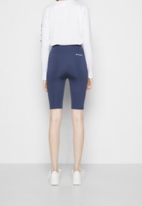 Personne portant un cuissard de vélo Columbia bleu marine et une chemise blanche à manches longues, vue de dos, debout sur un fond uni.