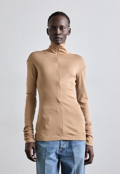 Haut beige à manches longues avec un col montant, fabriqué dans un tissu lisse, présentant un détail de couture sur le devant et un design ajusté. Associé à un jean bleu.