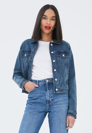 Frau mit glatten dunklen Haaren, die eine blaue Jeansjacke, ein weißes T-Shirt und hochtaillierte blaue Jeans trägt, posiert mit einer Hand in der Tasche.