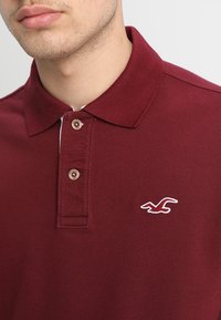 Burgundská polo košile vyrobená z texturované bavlny, s klasickým límečkem, dvěma knoflíky a bílým vyšívaným logem na levé straně hrudi.