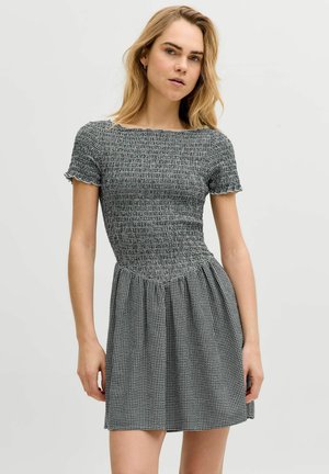 Jonge vrouw met blond haar die een zwart-witte, getextureerde, gesmockte jurk met geplooide rok draagt tegen een effen achtergrond.