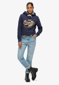 Sudadera con capucha azul marino y letras doradas, jeans de mezclilla azul claro y botas negras con cordones. Las mangas de una camiseta a rayas asoman por la capucha.