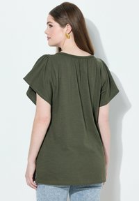 Oliivinvihreä lyhythihainen paita, jossa on pyöreä pääntie, takana rypytettyjä yksityiskohtia ja rento istuvuus, yhdistettynä vaaleisiin farkkuihin.