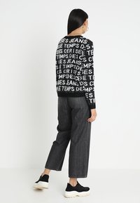 Pull en tricot noir et blanc avec motif de texte, associé à un jean large taille haute gris foncé. Des chaussures de sport noires complètent le look.