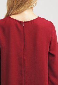 Abito bordeaux con scollatura rotonda, maniche lunghe e dettaglio con zip sul retro. Il tessuto ha un aspetto liscio e leggermente testurizzato.