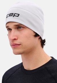 Lichtgrijze beanie van zacht stof, met een gestructureerd ontwerp en een zwart logo aan de voorkant. Draagbaar met een donker shirt.