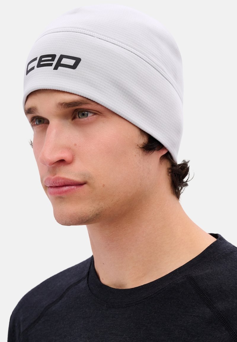 Lichtgrijze beanie van zacht stof, met een gestructureerd ontwerp en een zwart logo aan de voorkant. Draagbaar met een donker shirt.