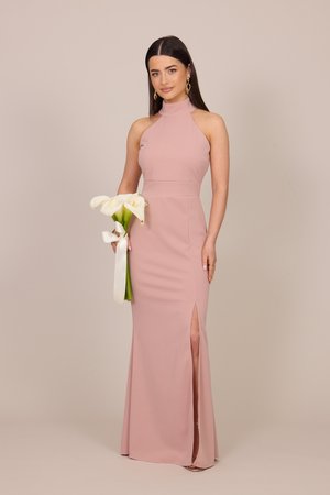 Kobieta w różowej sukni bez rękawów z wysokim rozcięciem, trzymająca białe kalii związane wstążką, stojąca na jednolitym beżowym tle.