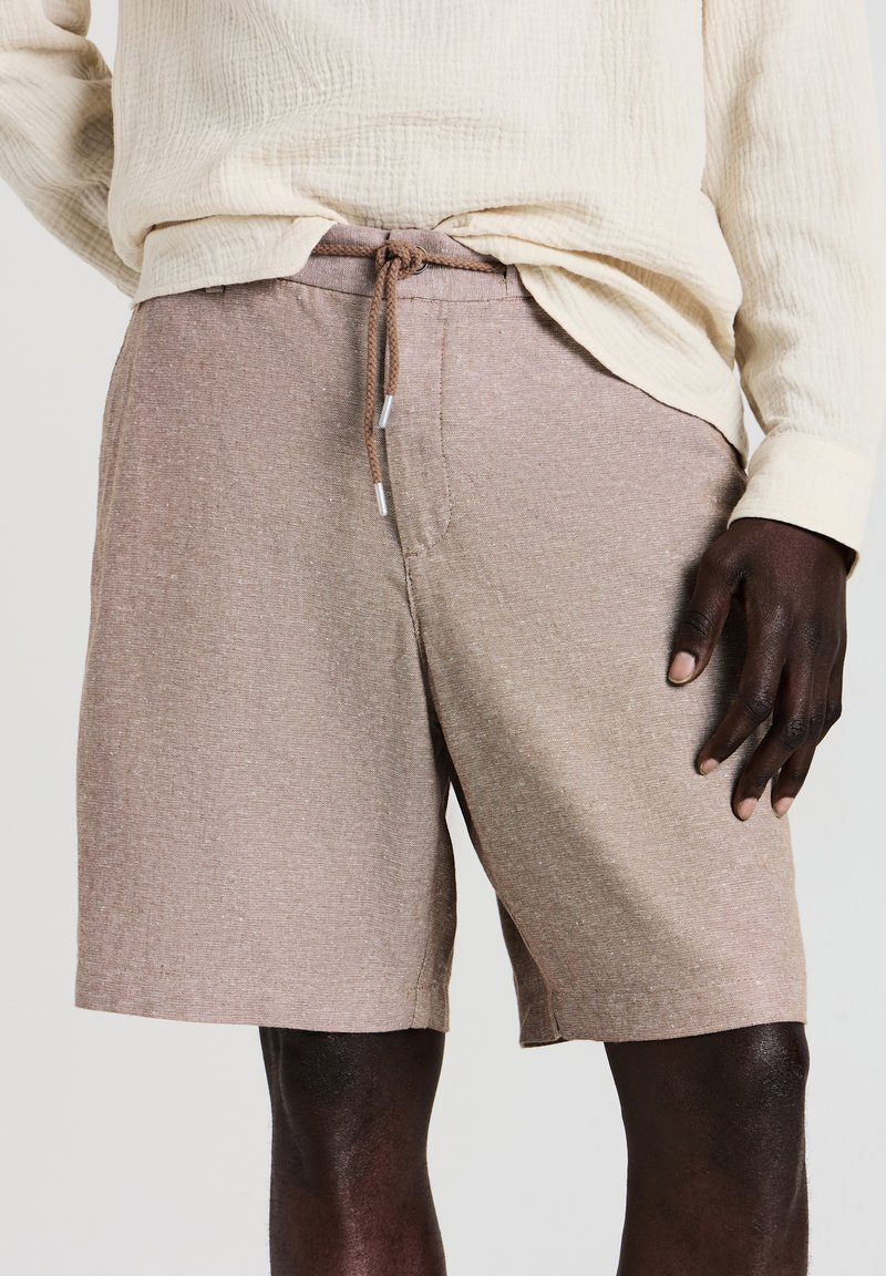 Beige strukturierte Shorts mit Kordelzug, getragen von einer Person mit dunkler Hautfarbe, kombiniert mit einem cremefarbenen Langarmshirt aus Baumwolle, das teilweise eingesteckt ist.