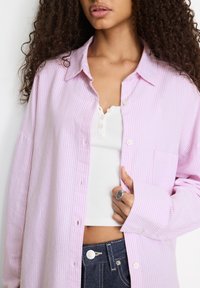 Camicia a righe rosa chiaro e bianca con maniche lunghe, una tasca sul petto e vestibilità comoda, indossata sopra una canottiera bianca.