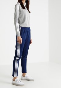 Grijze langwerpige top met drie witte strepen op de schouders, gecombineerd met marineblauwe tapered broek met drie witte zijstrepen, gedragen met witte sneakers.