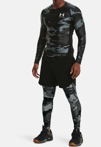 Man in zwart en grijs camouflage compressietop en leggings, zwarte atletische shorts en zwarte sportschoenen met rubberen zolen.