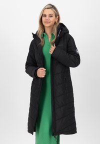 Cappotto imbottito nero con cappuccio, caratterizzato da un design trapuntato, chiusura frontale con zip e tasche laterali, indossato sopra un vestito verde a costine.
