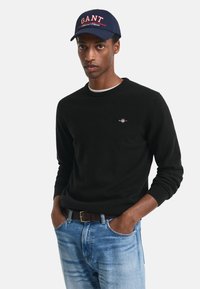 Maglione nero con colletto rotondo, indossato sopra una camicia bianca, abbinato a jeans denim blu. Cappellino navy con logo "GANT" in rosso.