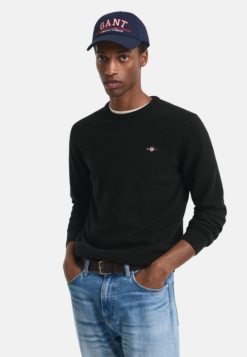 Maglione nero con colletto rotondo, indossato sopra una camicia bianca, abbinato a jeans denim blu. Cappellino navy con logo "GANT" in rosso.