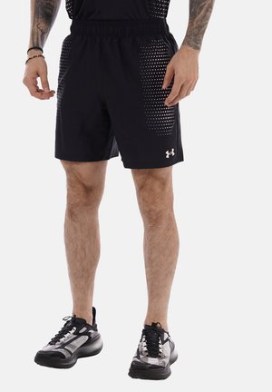 Uomo che indossa pantaloncini sportivi neri con motivi laterali bianchi a puntini e scarpe da corsa nere e bianche, in piedi davanti a uno sfondo uniforme.